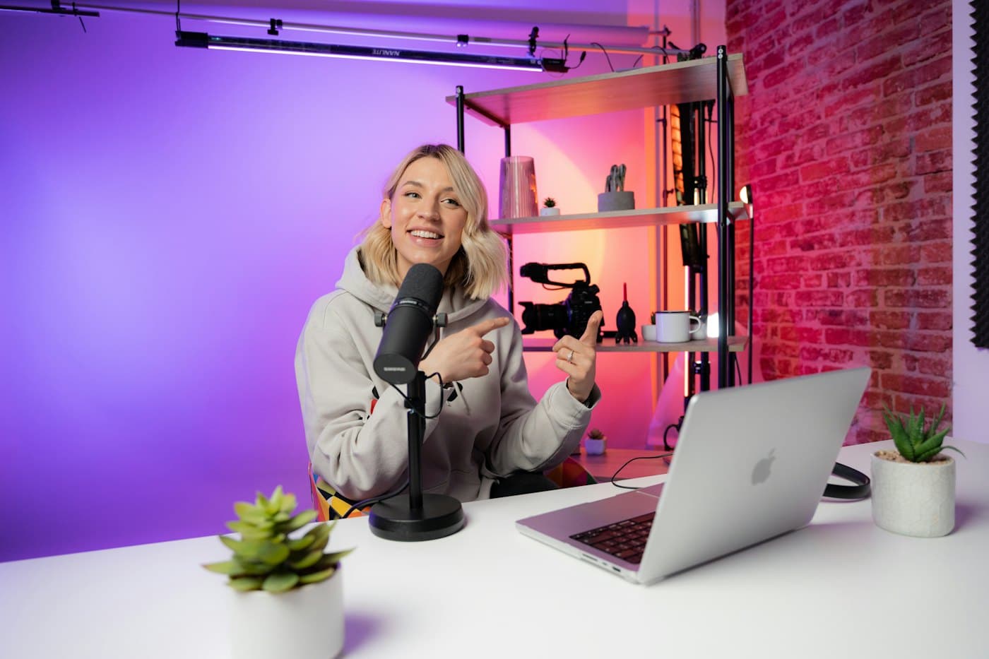A creator smiling at her desk with a microphone, laptop and camera in a studio lit pink and purple